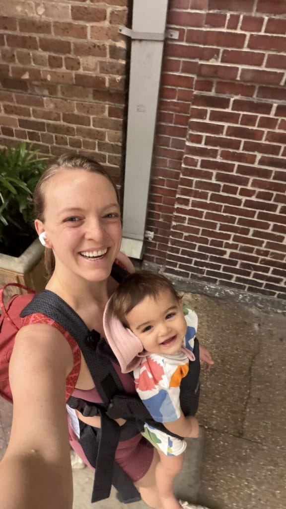 Emma Larson smiling for a selfie with her baby in a front carrier on a city sidewalk