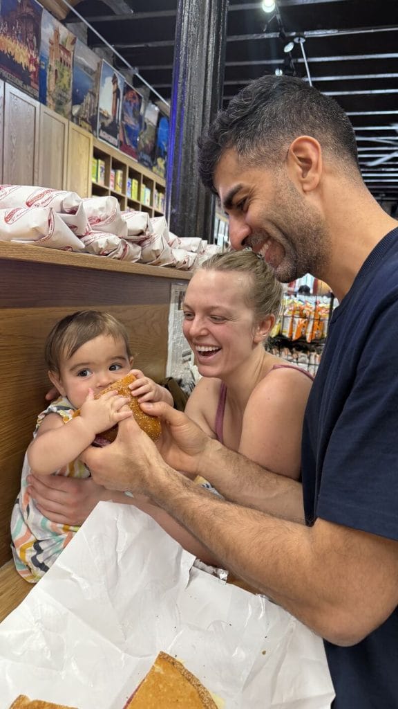 Emma Larson and her husband laughing as their baby bites into a sandwich at a New Orleans market
